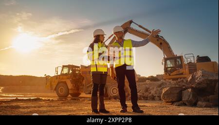 Caucasian Male Engineer Talking To Female Hispanic Architect With Laptop On Construction Site Of Apartment Building. Real Estate Developer Discussing Infrastructure. Heavy Machinery Working. Cinematic Stock Photo