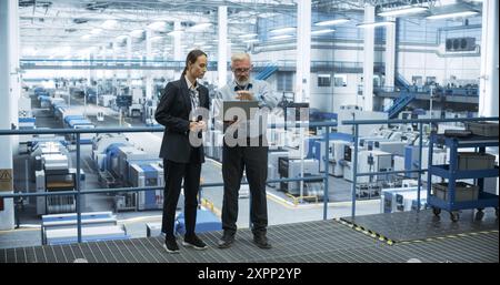 Middle Aged Engineer and Young Female Technician Having a Conversation and Using Laptop Computer. Specialists Monitoring, Analyzing System Reports at a Modern Electronics Factory Stock Photo