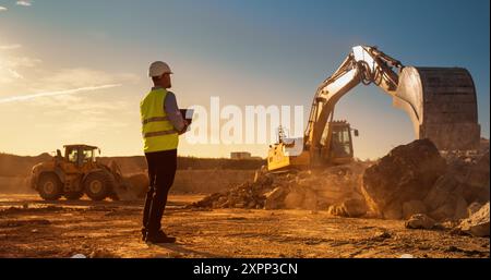 Caucasian Male Inspector Using Laptop Computer On Construction Site Of New Real Estate Project. Man Observing Industrial Machinery At Work, Laying Foundation For Apartment Building. Sunny Evening Stock Photo