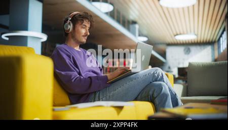 Portrait of a Smart Young Man Wearing Headphones and Working on a Laptop Computer in a Library Lobby. Young Man Sitting Comfortably on a Couch, Doing a Homework Assignment and Preparing for Exams Stock Photo