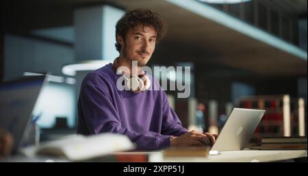Portrait of a Handsome European Student Studying in a Traditional Library. Young Male Looking at Camera and Smiling. Scholar Working on Laptop Computer in College, Researching Academic Topics Online Stock Photo
