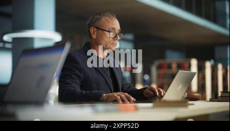 Knowledgeable Teacher Using Laptop Computer in a Library. Smart Middle Aged Male Educator Preparing Lectures for College Students, Checking Exams, Browsing and Researching Internet Articles Stock Photo