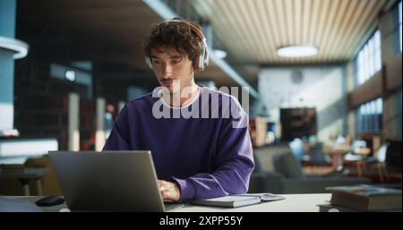 Handsome Student Writing an Essay on Laptop Computer, Thinking About the Topic, Brainstorming, Finding Solutions. Young Stylish Man Studying in a Public Library, Listening to Lecture in Headphones Stock Photo