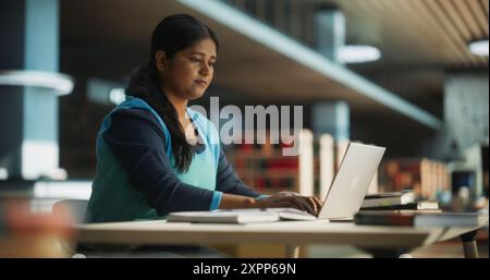 Focused Indian Female Working on a College Project for Class Assignment, Using Online Office Software on Laptop Computer in a Public Library Space. Young South Asian Woman Learning New Skills Online Stock Photo