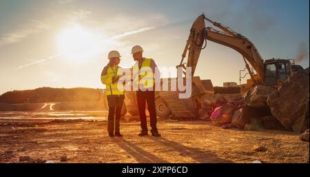 Hispanic Female Urban Planner Using Laptop And Talking To Caucasian Male Real Estate Investor On Construction Site. Colleagues Discussing New Project While Heavy Machinery Working. Warm Edit Stock Photo