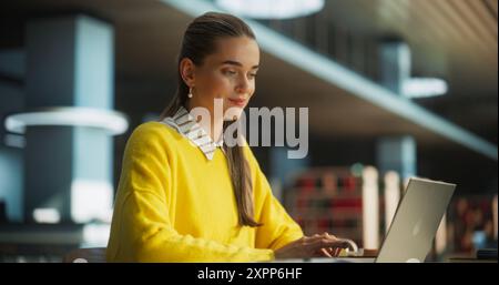 Excited Young Female is Focused on Creating an University Presentation for Class Assignment, Using Laptop Computer and Taking Notes in a Notebook in a Public Library Space Stock Photo