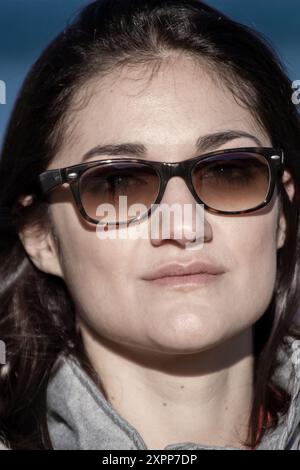 A woman wearing sunglasses and a gray jacket. She has a serious expression on her face. sunglasses are black and have a brown tint Stock Photo