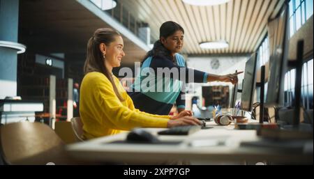 Two Multiethnic Girlfriends Studying in an University Library. Smart Indian Female Helping Her Classmate Colleague with Homework. Couple Using Desktop Computer for a Research and Exam Preparation Stock Photo