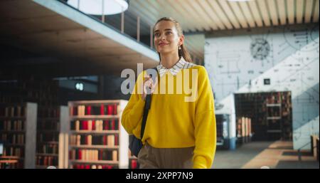 Beautiful Caucasian Female Student Walking in an Empty Public Library with Bookshelves. Young Smart Woman Carrying a Backpack with Textbooks to Prepare and Study for Exams. Low Angle Portrait Stock Photo