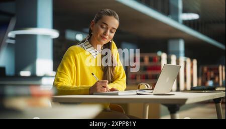 Thoughtful European College Student Using Laptop Computer to Study in a Public Library. Beautiful Young Woman Learning Online, Getting Ready for Exams, Taking Notes and Drafting an Essay Stock Photo