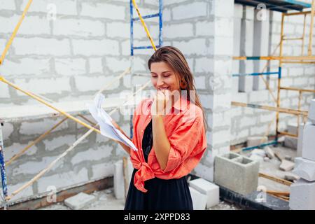 Portrait of happy young woman checks her house under construction holding project blueprint plan on paper. Future owner imagines cozy home standing by Stock Photo