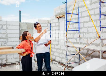 Man architect engineer showing house under construction to woman future homeowner checking blueprint plan. Consulting during project while building wi Stock Photo