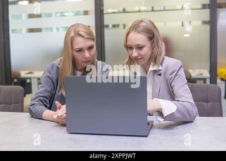 Two young businesswomen working on laptop in workspace. They are sitting at table and looking at laptop screen together. Stock Photo