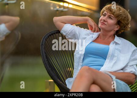 A woman in her mid-40s sits on the terrace and gazes with a dreamy eye towards the side. Stock Photo