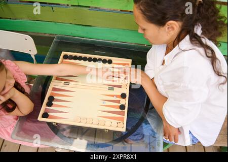 Two children engaged in a fun game of backgammon, playing together outside on a vibrant, colorful wooden table. Perfect for themes of family time, pla Stock Photo