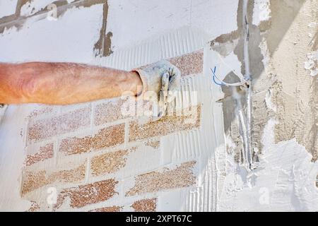 External cladding of facade house with thin faux brick panels, construction worker uses tile adhesive. Stock Photo