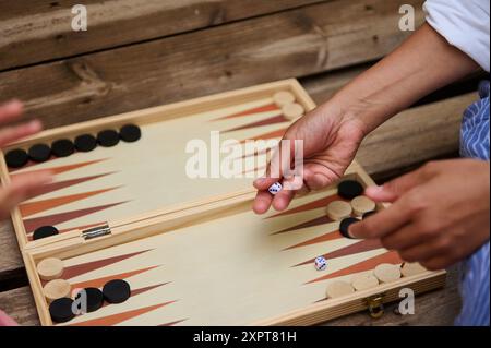 A close-up shot of friends enjoying a game of backgammon on a wooden table outdoors. The image captures the hands of individuals rolling dice and movi Stock Photo