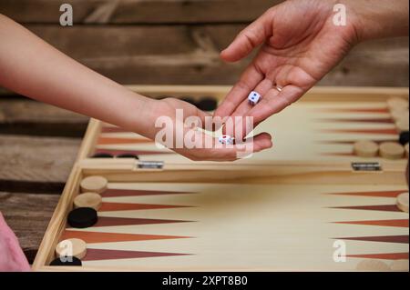 Close-up of adult and child hands passing dice while playing backgammon on a wooden table. Concept of family bonding, leisure, and traditional games. Stock Photo