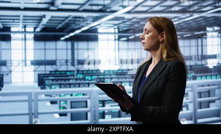 Meticulous technician doing checkup, inputting data on tablet of needed fixes in server hub housing advanced data storage infrastructure supporting critical IT workloads, camera B Stock Photo