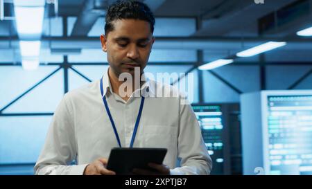 Programmer walking in high tech data center used for managing infrastructure energy consumption, working on tablet. Indian man using device in workspace used for automation in server farm, camera A Stock Photo