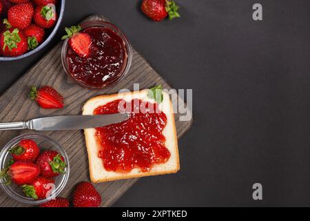 Slice of bread toast with strawberry jam, fresh strawberries in bowls, and a knife on a dark background. Stock Photo