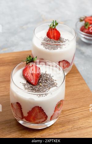Two glasses of creamy yogurt topped with fresh strawberry halves and chia seeds on wooden surface, healthy breakfast Stock Photo