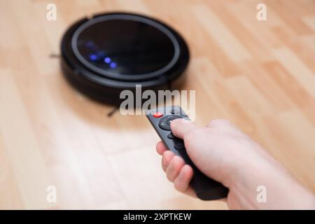 Hand holding remote control, operating a robot vacuum on a wooden floor,smart home technology, hygiene Stock Photo