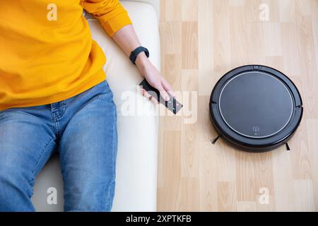 Man in yellow sweater holding a remote control while sitting on a white sofa, directing a robot vacuum cleaner on a wooden floor. Stock Photo