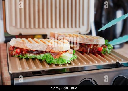 Two grilled sandwiches with lettuce, bacon, and tomatoes cooking on a panini press in a kitchen Stock Photo