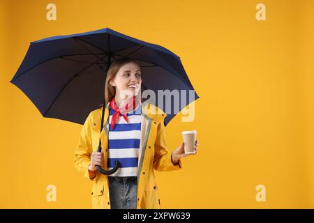 Woman with blue umbrella and paper cup on yellow background, space for text Stock Photo