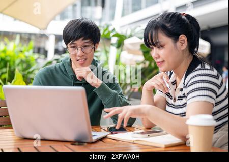 Two young Asian college students are looking at a laptop screen, discussing work and doing on a co-project together at a cafe in the city. Stock Photo