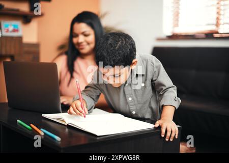 Book, writing and child with homework in house with mother working on laptop for kindergarten education. Learning, drawing and boy kid student with Stock Photo