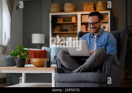 Smiling male web designers dressed in denim shirt working over laptop and sitting with crossed legs on armchair. Young businessman coding over wireles Stock Photo