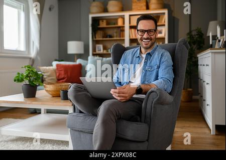 Portrait of confident happy male professional with laptop sitting on armchair in home office. Smiling young web designer coding over wireless computer Stock Photo