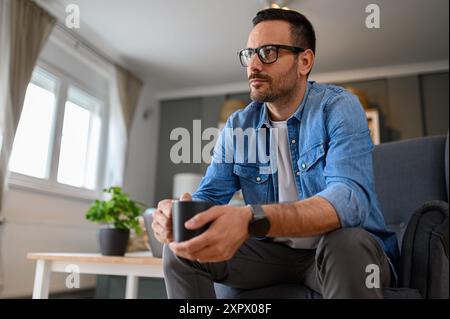 Low angle view of serious young businessman holding coffee cup and contemplating while sitting on armchair. Male freelancer having drink and thinking Stock Photo