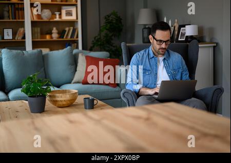 Serious young businessman wearing eyeglasses sending business e-mails over laptop while sitting on armchair. Confident male freelancer analyzing data Stock Photo