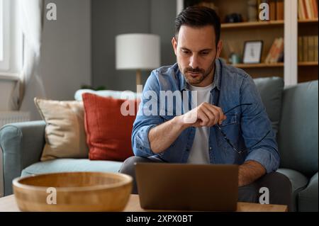 Focused male computer programmer holding eyeglasses and working over laptop seriously while sitting on sofa. Young pensive freelancer coding over wire Stock Photo