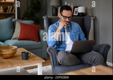 Serious focused male freelancer with hand on chin thinking and analyzing data over laptop while sitting on armchair. Young businessman coding over wir Stock Photo