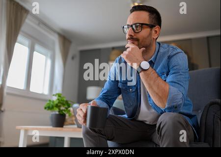 Positive male entrepreneur with hand on chin thinking business strategies while sitting comfortably on armchair. Young pensive freelancer holding coff Stock Photo