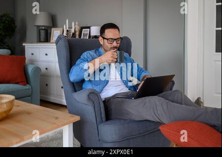 Male entrepreneur drinking coffee and making business plans over wireless computer while relaxing on armchair. Young handsome freelancer working remot Stock Photo