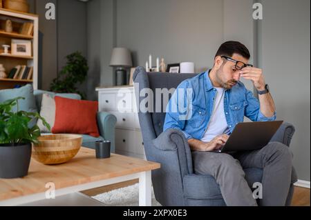 Exhausted young businessman touching head in pain and checking project deadlines over laptop while sitting on armchair. Stressed male freelancer worki Stock Photo