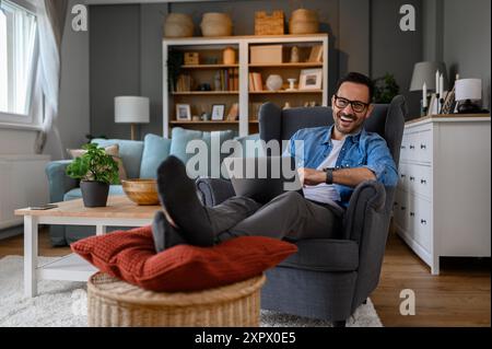 Portrait of cheerful young businessman laughing and doing research over laptop while relaxing legs on footstool. Male entrepreneur working over comput Stock Photo