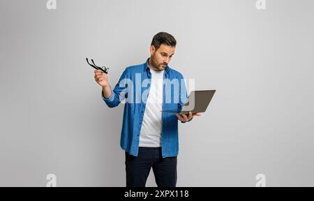 Shocked young businessman holding glasses and looking at laptop while standing on white background Stock Photo