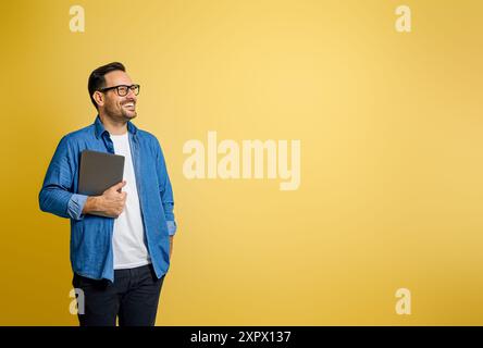 Happy confident male entrepreneur holding digital tablet and contemplating over yellow background Stock Photo