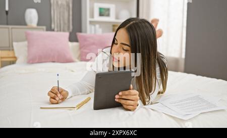 A focused young hispanic woman writes in a notebook and uses a tablet in her well-lit bedroom. Stock Photo