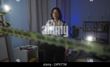 A mature hispanic woman detective examines an indoor crime scene with flashlight, caution tape, and evidence. Stock Photo