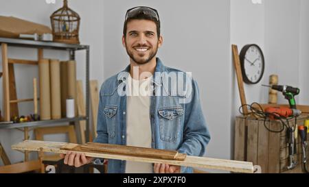 Handsome young man in denim jacket holding lumber with carpentry tools in a woodshop. Stock Photo