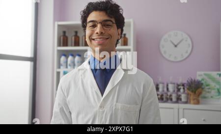 A young hispanic man in a pharmacy shop wearing a lab coat with a friendly smile and glasses, standing indoors with shelves and products visible in th Stock Photo