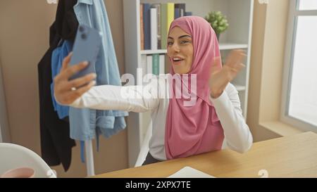 Smiling woman wearing hijab taking selfie indoors with smartphone in a bright room Stock Photo