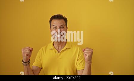 Hispanic man celebrating with a wide smile and clenched fists, dressed in a yellow shirt, standing in front of a bright yellow background. Stock Photo